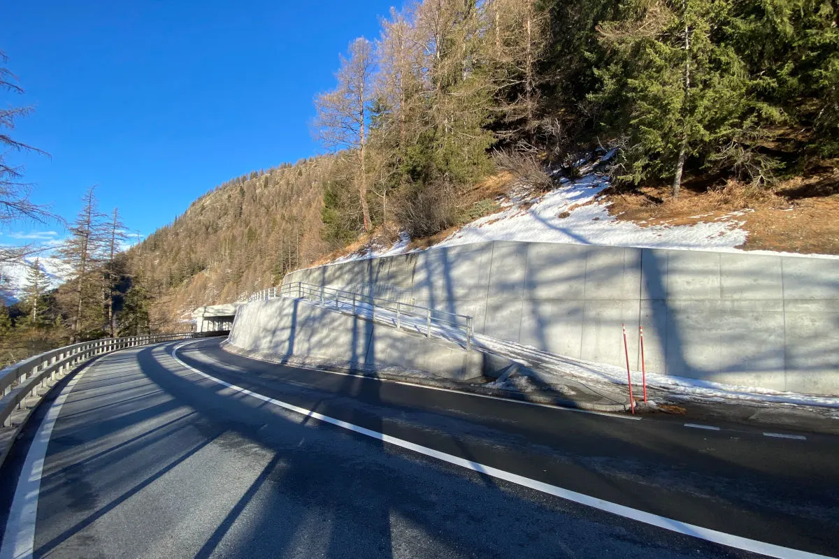 Das Bild zeigt eine leere Bergstraße mit einer Kurve. Eine Betonstützmauer ist rechts zu sehen, daneben ein schneebedeckter Hang.
