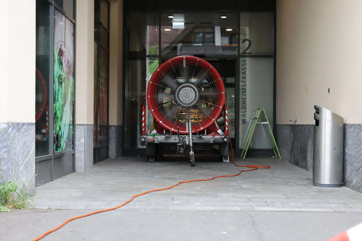 Ein großer, roter mobiler Ventilator steht in einer überdachten Durchfahrt eines Gebäudes. Ein Stromkabel ist angeschlossen und verläuft über den Boden.