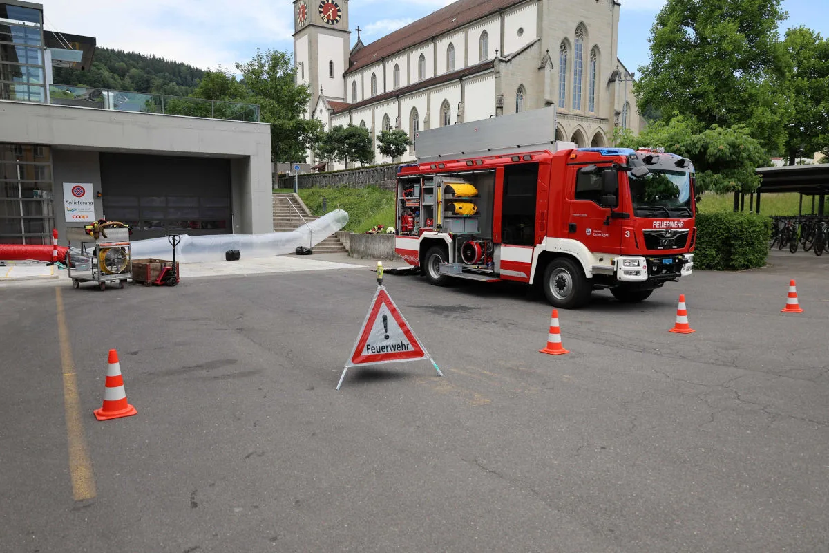 Ein rotes Feuerwehrfahrzeug mit geöffnetem Geräteraum steht auf einem Parkplatz. Im Hintergrund ist eine Kirche sichtbar.