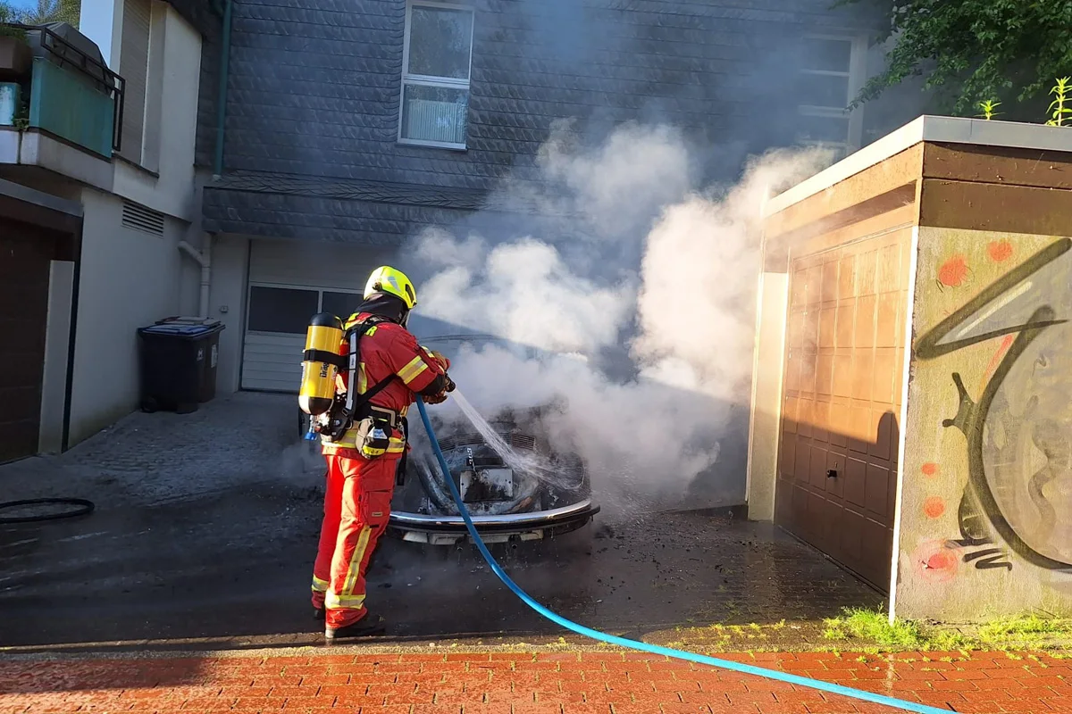 Ein Feuerwehrmann in voller Ausrüstung löscht ein brennendes Fahrzeug in einer Einfahrt. Dicker Rauch steigt auf, während er den Wasserschlauch bedient.