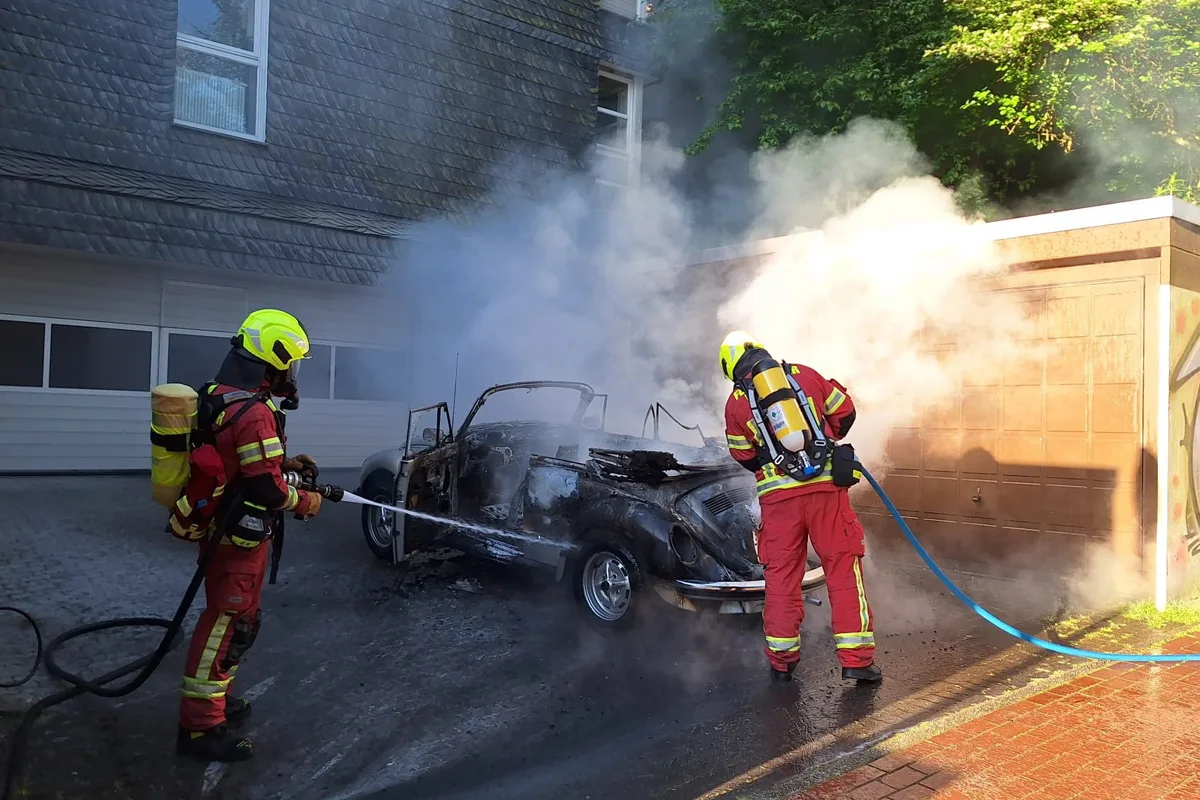 Zwei Feuerwehrleute in Schutzkleidung löschen ein brennendes Auto vor einer Garage in einer Wohngegend.