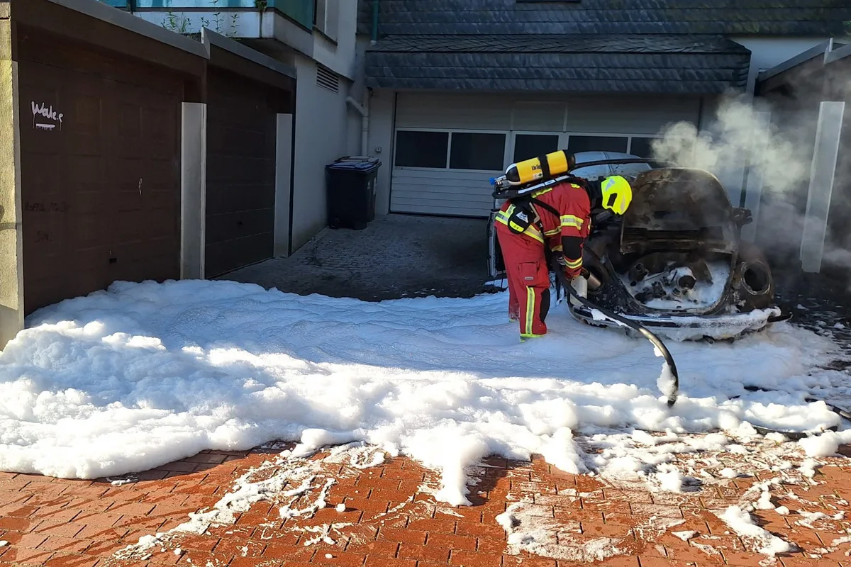 Ein Feuerwehrmann in roter Uniform mit Atemschutz löscht einen ausgebrannten PKW vor einer Garage mit Schaum. Der Boden ist mit Löschschaum bedeckt.