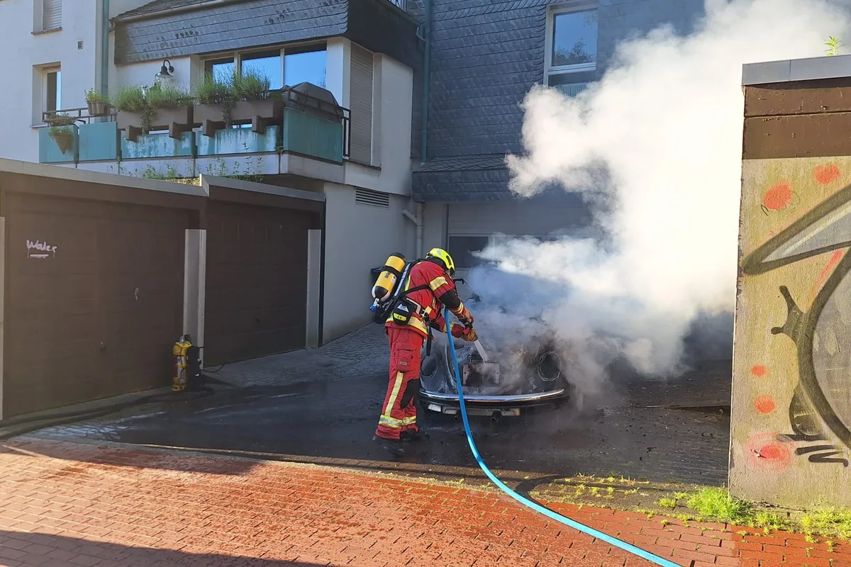 Ein Feuerwehrmann, ausgerüstet mit Atemschutz, bekämpft einen Fahrzeugbrand in einer Einfahrt, umgeben von dichter Rauchentwicklung.