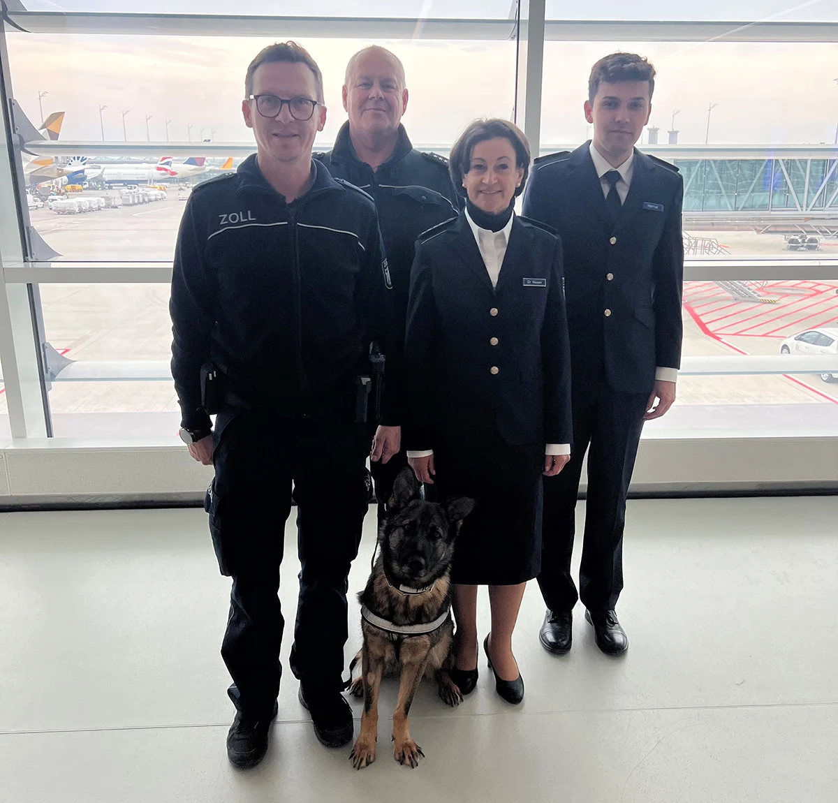 Vier Zollbeamte in Uniform stehen neben einem Diensthund in einem hellen Flughafenterminal. Im Hintergrund ist das Rollfeld sichtbar.
