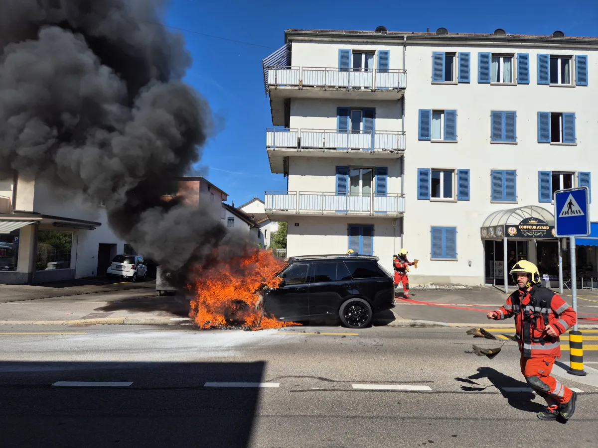 Auf einer Straße steht ein Auto in Flammen und es ist von dichtem Rauch umgeben. Feuerwehrleute in Uniform sind dabei, den Brand zu bekämpfen.