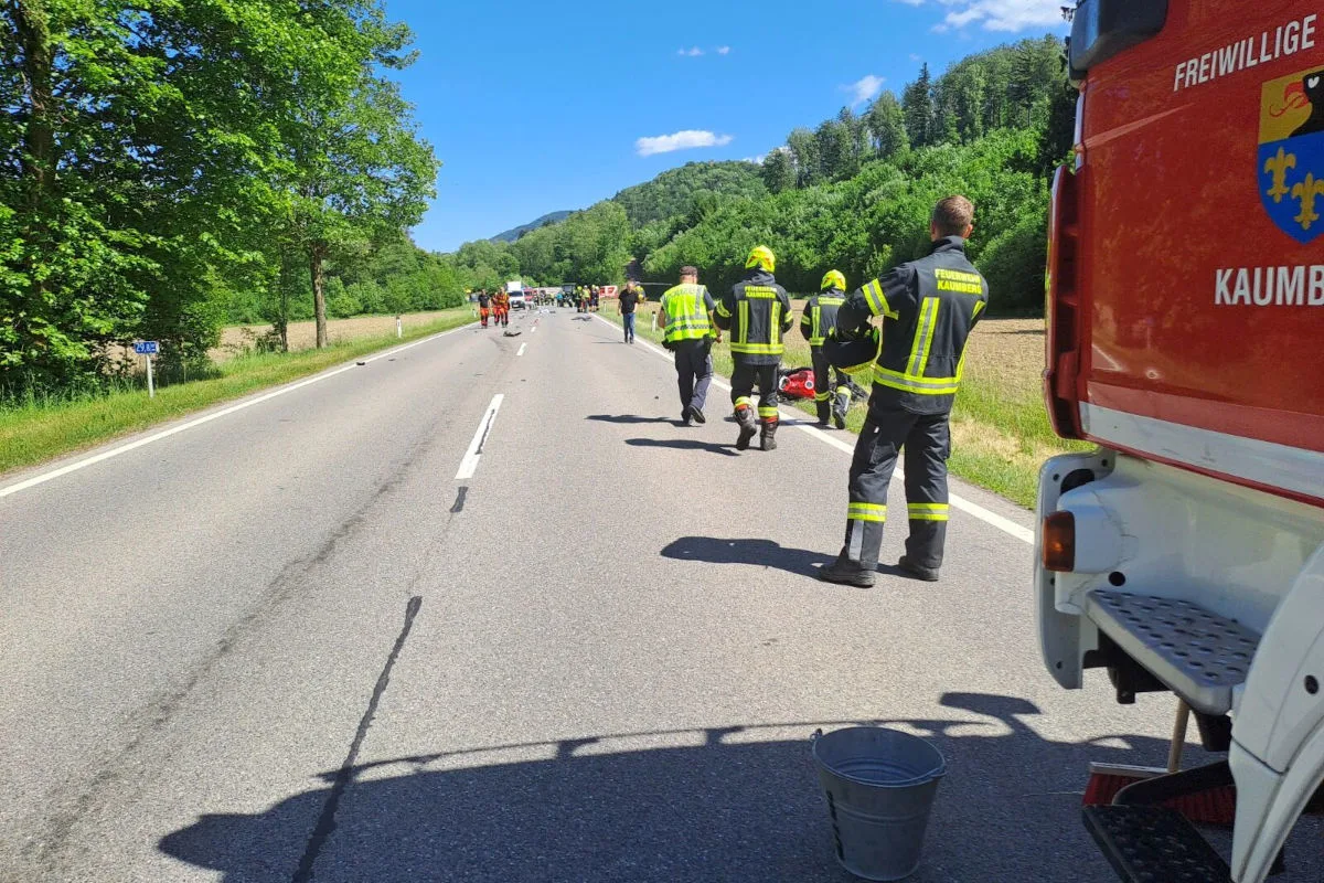 Mehrere Feuerwehrleute sind auf einer Landstraße in einer ländlichen Gegend bei strahlendem Sonnenschein im Einsatz. Im Vordergrund steht ein Feuerwehrfahrzeug der Freiwilligen Feuerwehr Kaumberg.