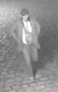 Eine Person geht nachts über Kopfsteinpflaster, aufgenommen von einer Überwachungskamera in Schwarz-Weiß.