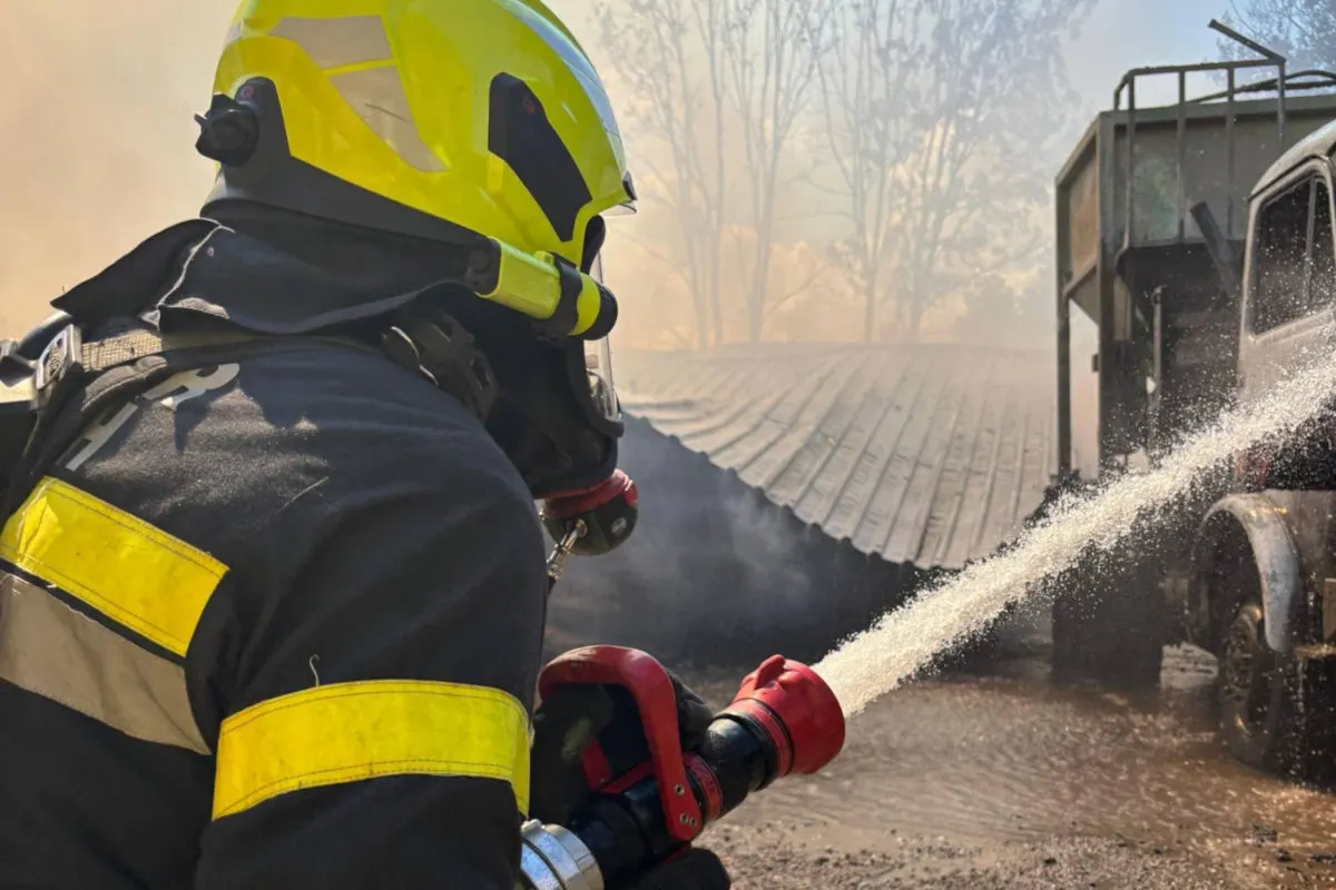 Ein Feuerwehrmann in Schutzkleidung und gelbem Helm löscht mit einem Wasserschlauch ein brennendes Fahrzeug. Im Hintergrund sind Bäume und Rauch sichtbar.