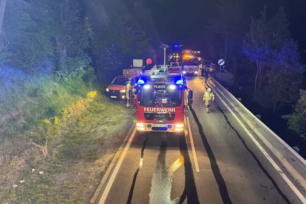 Ein rotes Feuerwehrfahrzeug mit blinkendem Blaulicht steht auf einer Landstraße. Zwei Feuerwehrleute sind in Schutzkleidung sichtbar.