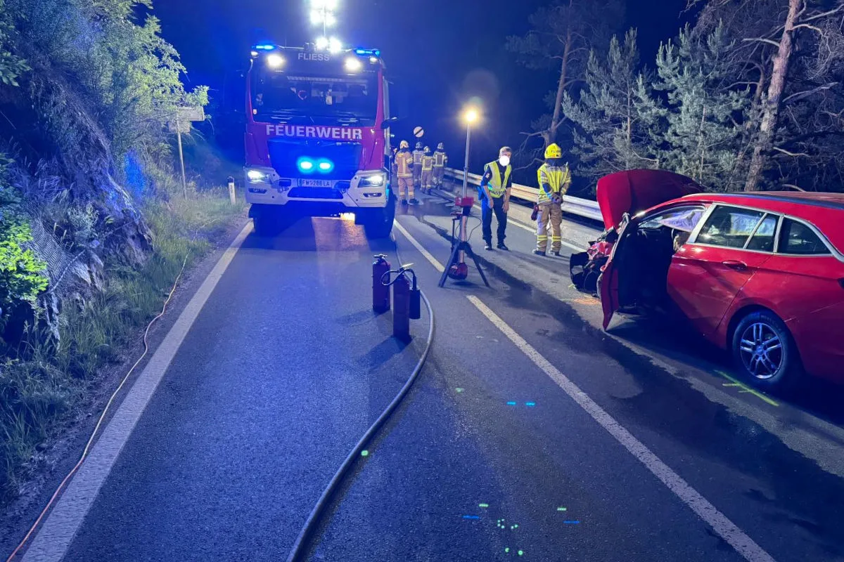 Ein rotes Feuerwehrfahrzeug mit Blaulicht steht auf einer Straße. Einsatzkräfte sind neben einem beschädigten roten Auto aktiv.