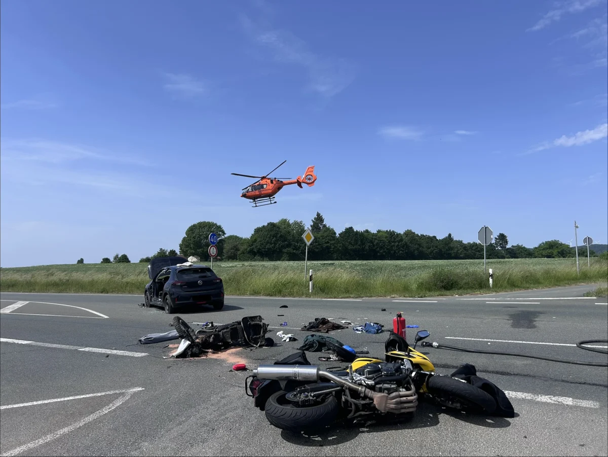 Ein Rettungshubschrauber fliegt über einer Unfallstelle, auf der ein beschädigtes Motorrad auf der Straße liegt. Im Hintergrund sind Felder und Bäume zu sehen.