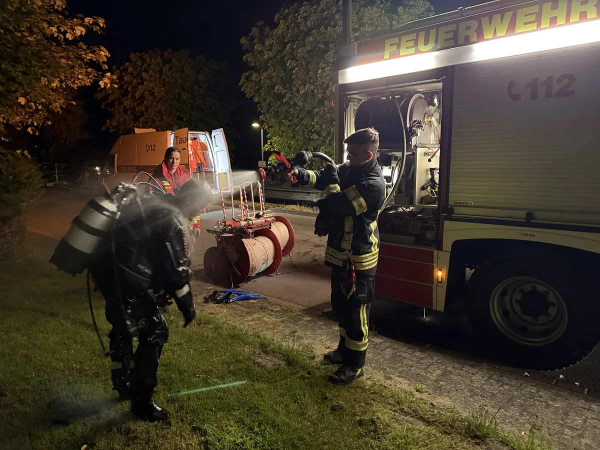 Ein Feuerwehrmann besprüht einen in Schutzkleidung gehüllten Kollegen mit Wasser. Im Hintergrund steht ein geöffnetes Feuerwehrfahrzeug bei Nacht.