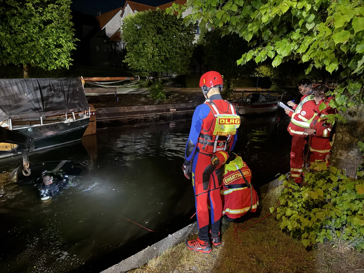 DLRG-Einsatzkräfte stehen am Ufer eines Kanals, während ein Taucher im Wasser nach Hinweisen sucht. Es ist Nacht, und der Bereich ist beleuchtet.