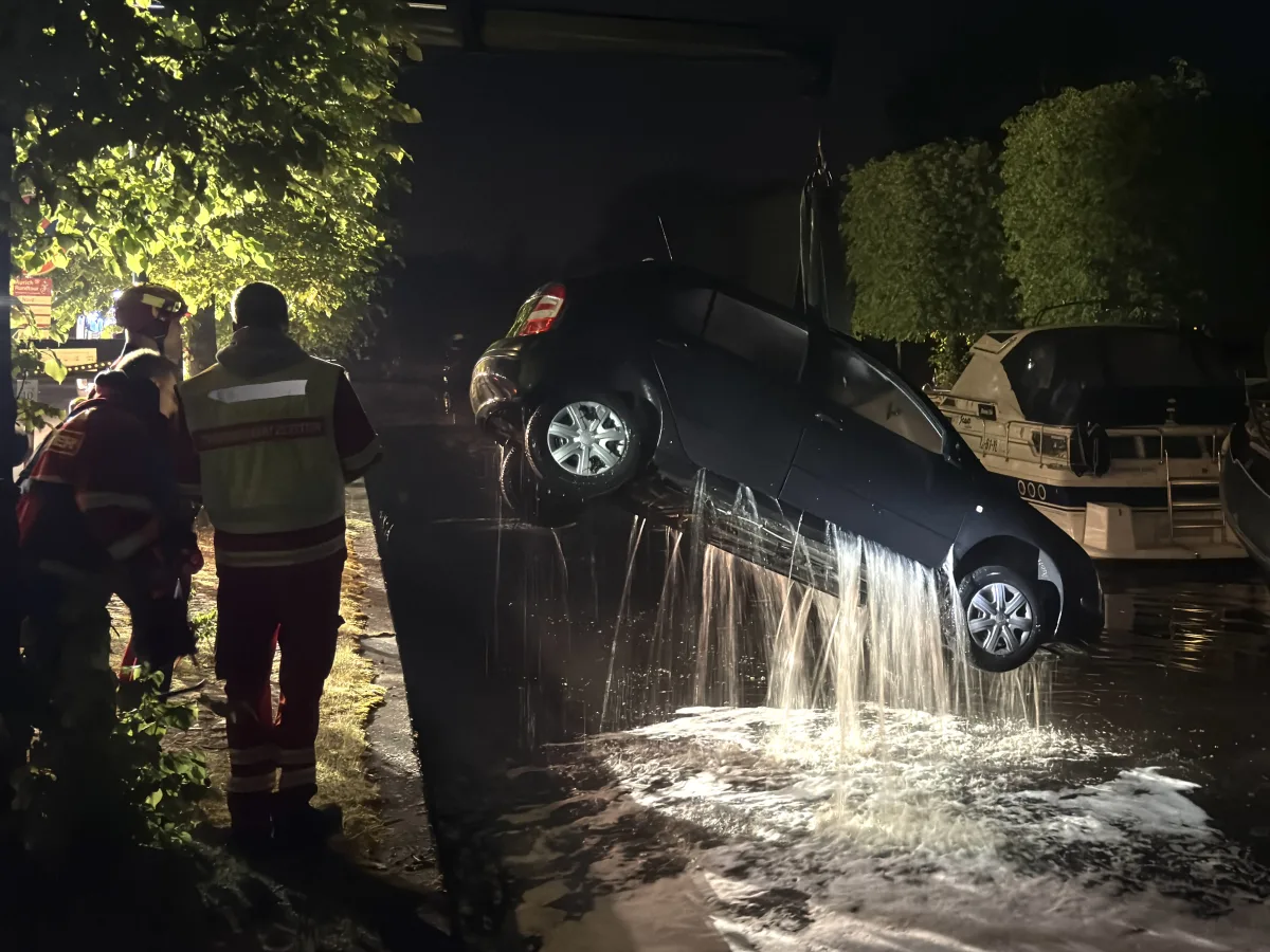 In der Nacht wird ein Auto aus dem Wasser gehoben, wobei Wasser aus dem Fahrzeug fließt. Feuerwehrleute sehen zu.