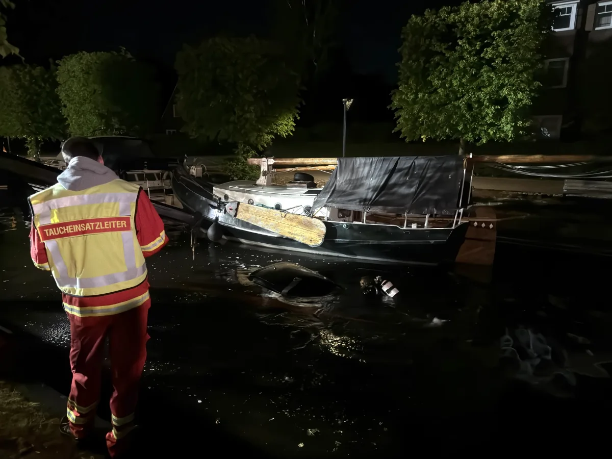 Ein Taucheinsatzleiter steht am Ufer und beobachtet, wie ein Taucher bei Nacht neben einem teilweise untergetauchten Boot arbeitet.