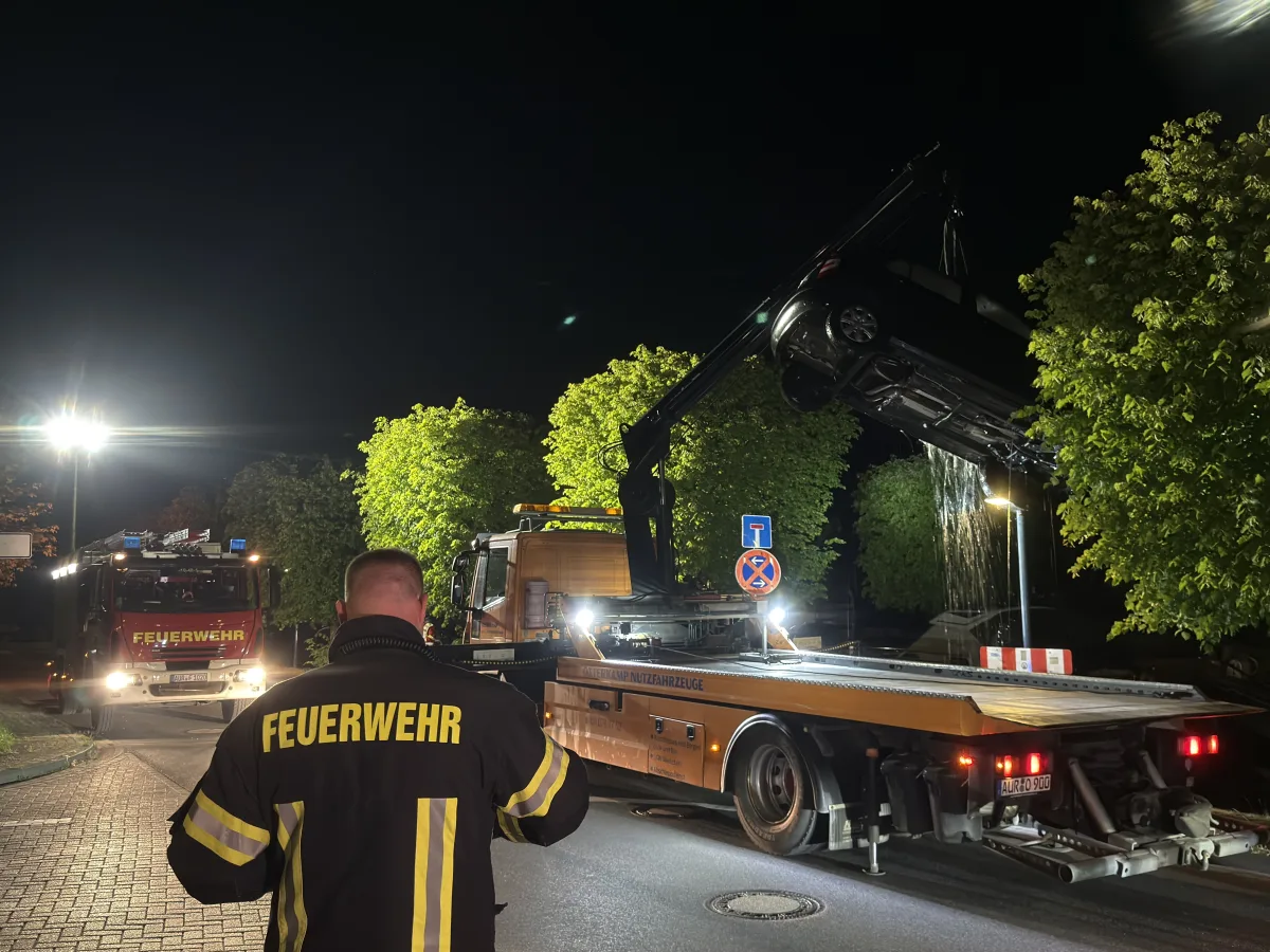 Ein Feuerwehrmann im Vordergrund beobachtet die nächtliche Bergung eines Fahrzeugs mithilfe eines Abschleppwagens. Im Hintergrund sind weitere Einsatzfahrzeuge zu sehen.
