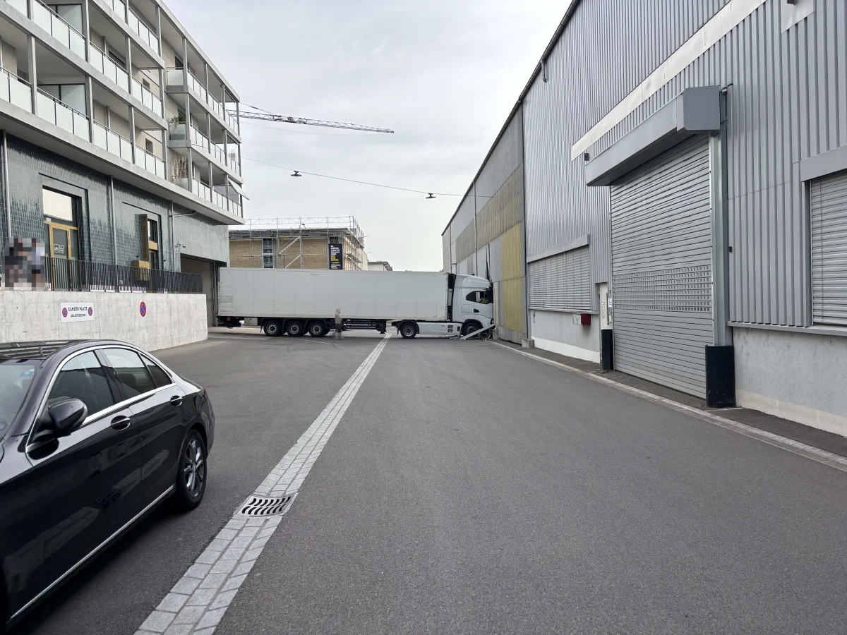 Ein großer weißer LKW steht quer in einem engen Industriehof. Links ein modernes Gebäude, rechts Industriehallen.