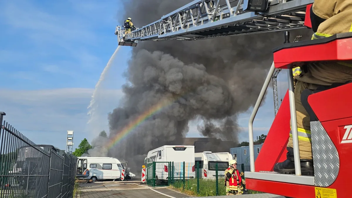 Ein Feuerwehrmann ist auf einer Leiter und löscht einen Brand, während dichter Rauch aufsteigt. Im Hintergrund sind Wohnmobile und ein Regenbogen sichtbar.