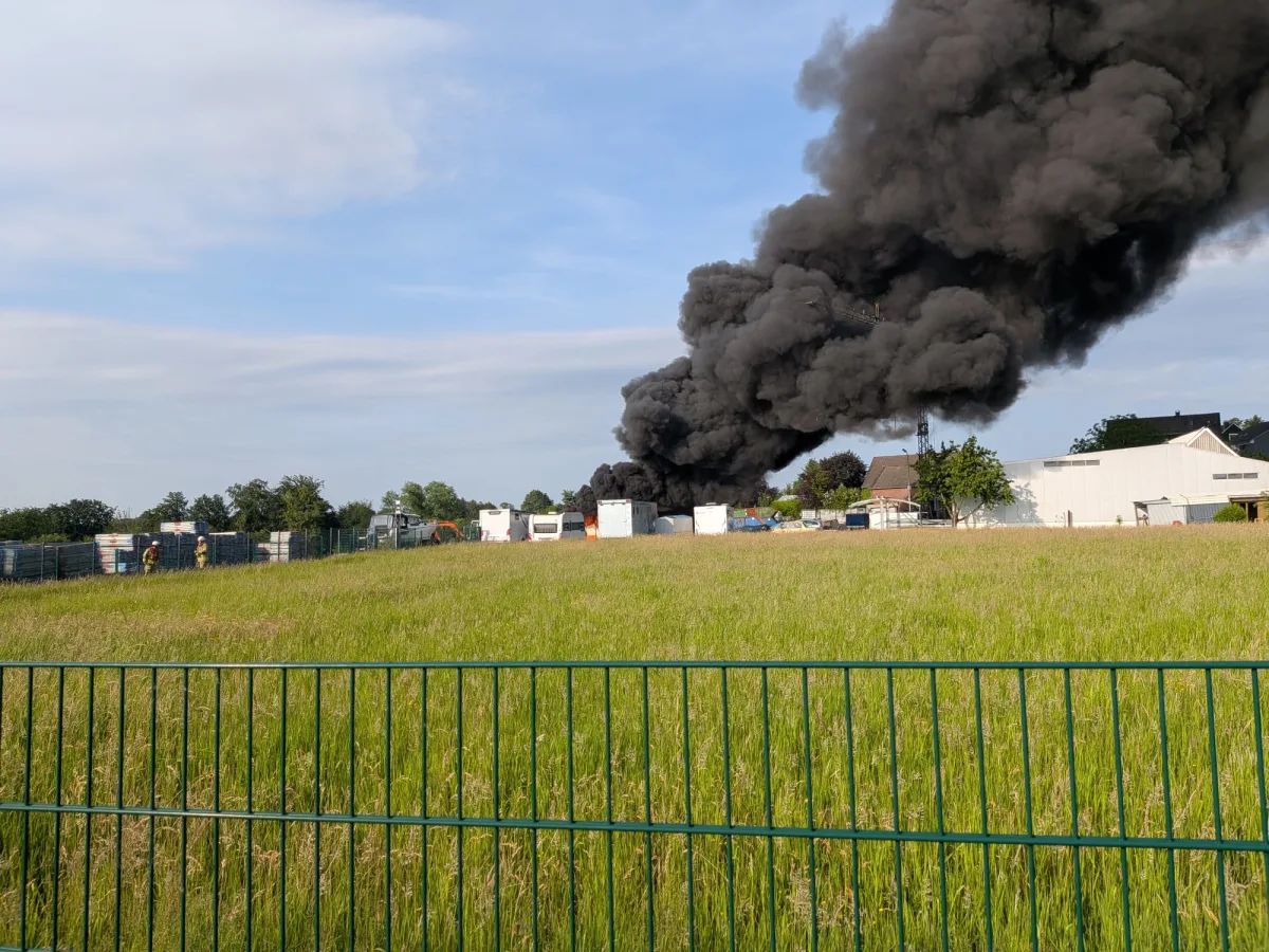 Dichte schwarze Rauchwolken steigen aus einem Gebäudekomplex in einem Industriegebiet auf. Im Vordergrund ist eine grüne Wiese zu sehen.