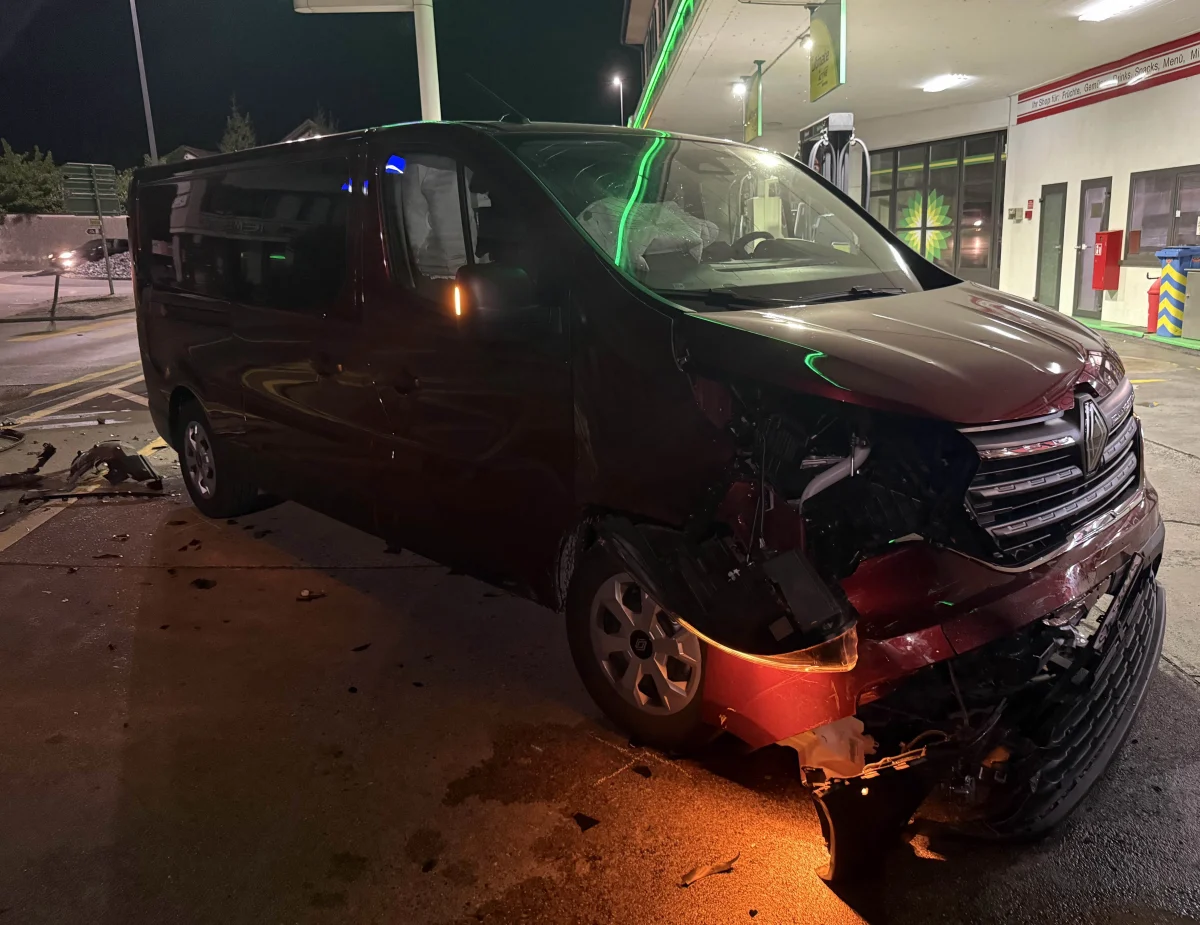 Ein dunkelroter Van mit erheblichem Frontschaden steht bei Nacht an einer beleuchteten Tankstelle. Auf dem Boden liegen Trümmerteile.