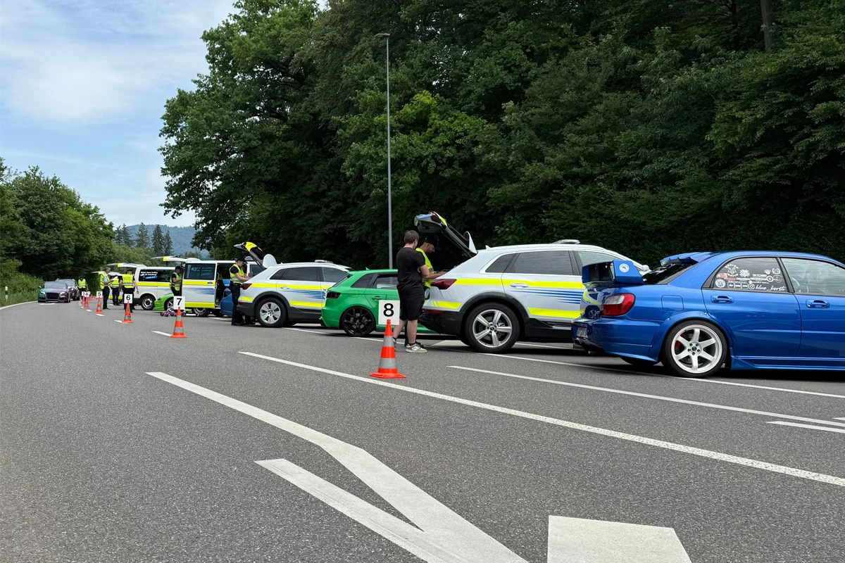 Autos stehen am Straßenrand einer Landstraße, darunter ein Polizeifahrzeug. Personen befinden sich bei den Fahrzeugen, Kofferräume sind geöffnet.