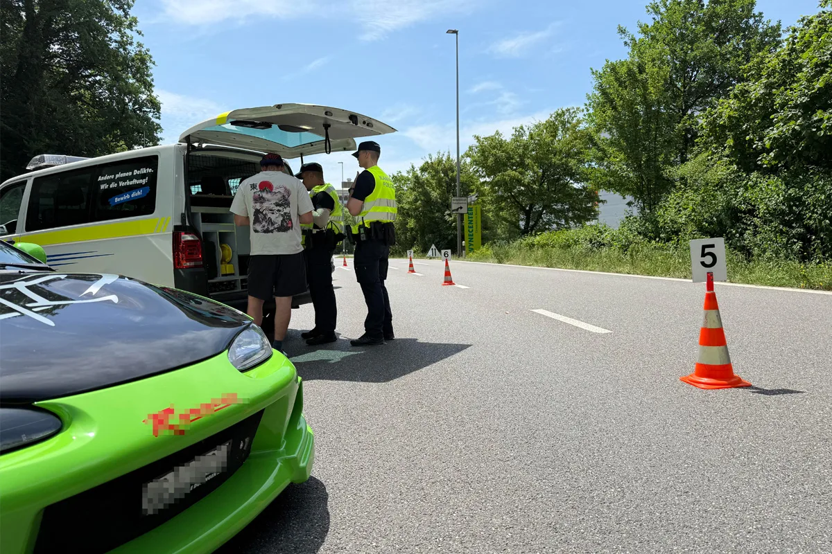 Ein grünes Auto steht neben einem Einsatzfahrzeug, bei dem Polizisten in gelben Westen mit einem Mann sprechen. Daneben stehen Verkehrskegel auf der Straße.