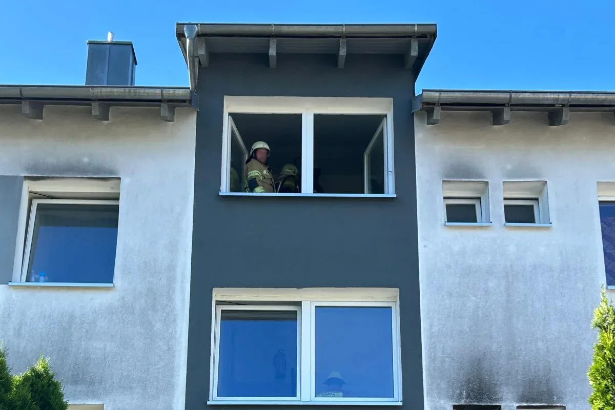 Mehrere Feuerwehrmänner stehen an einem geöffneten Fenster eines Gebäudes mit Rauchspuren an der Fassade.