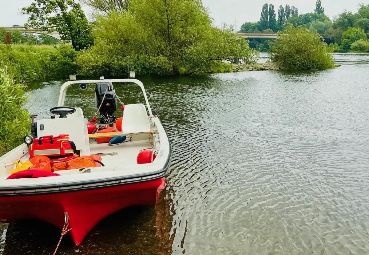 Ein rotes Rettungsboot liegt nahe am Ufer eines Flusses, umgeben von grünen Bäumen und ruhigem Wasser.