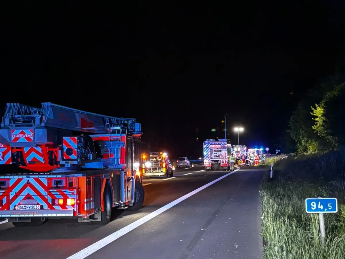 Feuerwehrfahrzeuge und Einsatzkräfte sind nachts auf einer beleuchteten Autobahn zu sehen. Mehrere Fahrzeuge stehen am Straßenrand.
