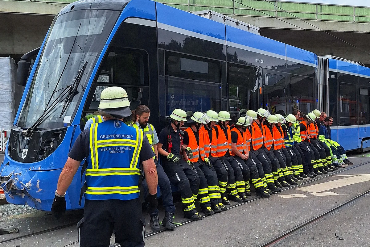 Mehrere Feuerwehrleute in orangenen Warnwesten sitzen auf einer blauen Straßenbahn vor Ort. Ein Gruppenführer beobachtet die Szene.