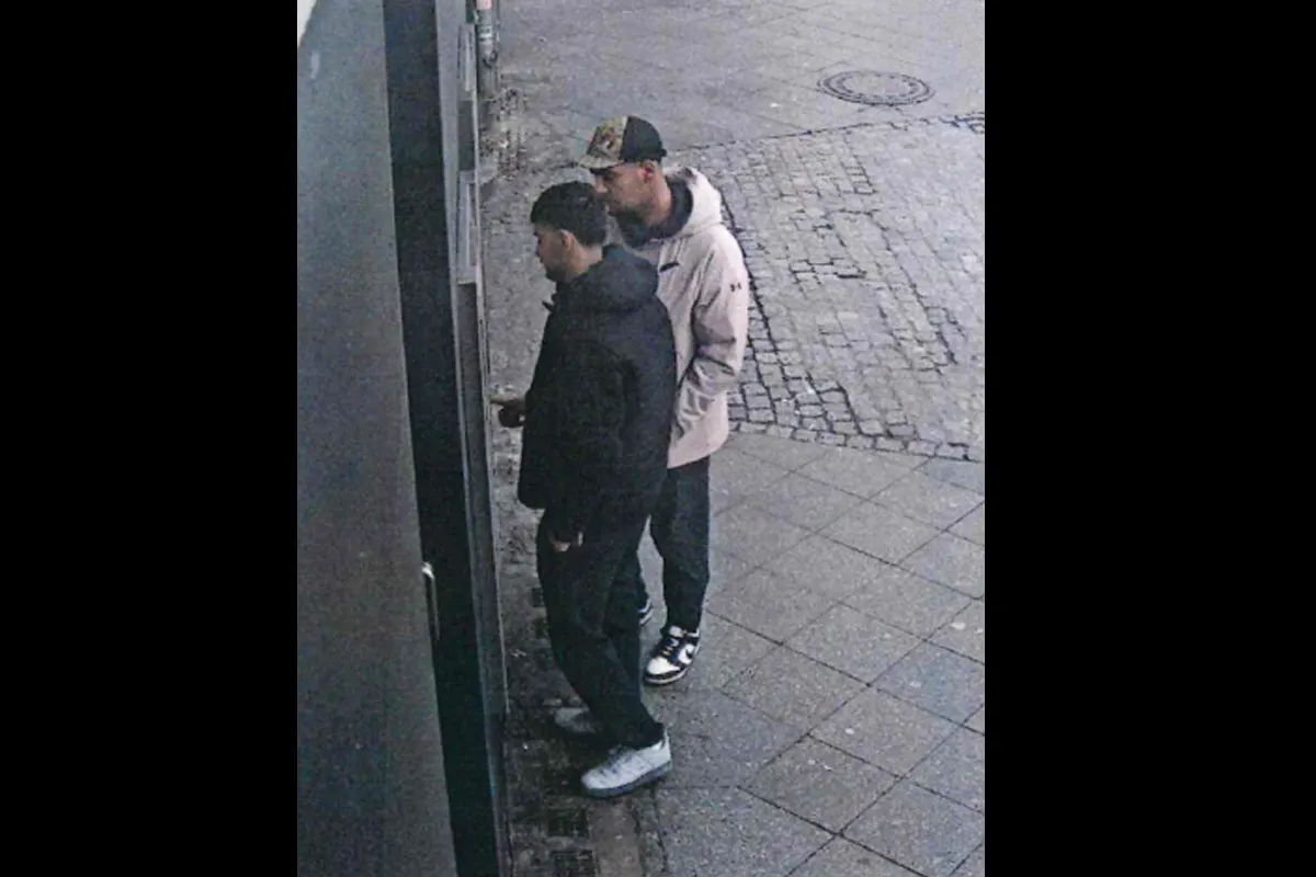 Zwei Männer stehen an einem Geldautomaten. Einer trägt eine helle Jacke und Kappe, der andere dunkle Kleidung. Der Boden ist gepflastert.