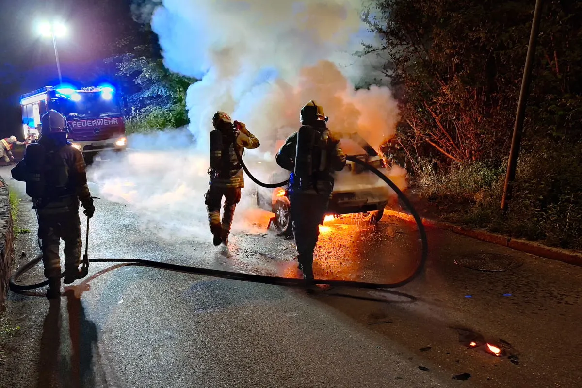 Drei Feuerwehrleute löschen mit Schläuchen einen brennenden PKW am Straßenrand bei Nacht. Flammen und Rauch steigen auf.