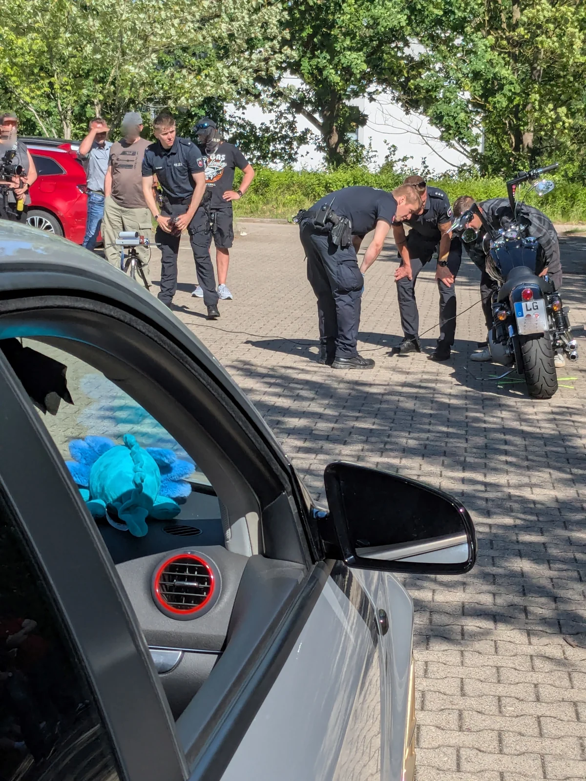 Mehrere Polizisten stehen zusammen auf einem Parkplatz und untersuchen ein Motorrad. Im Vordergrund ist ein Auto zu sehen.