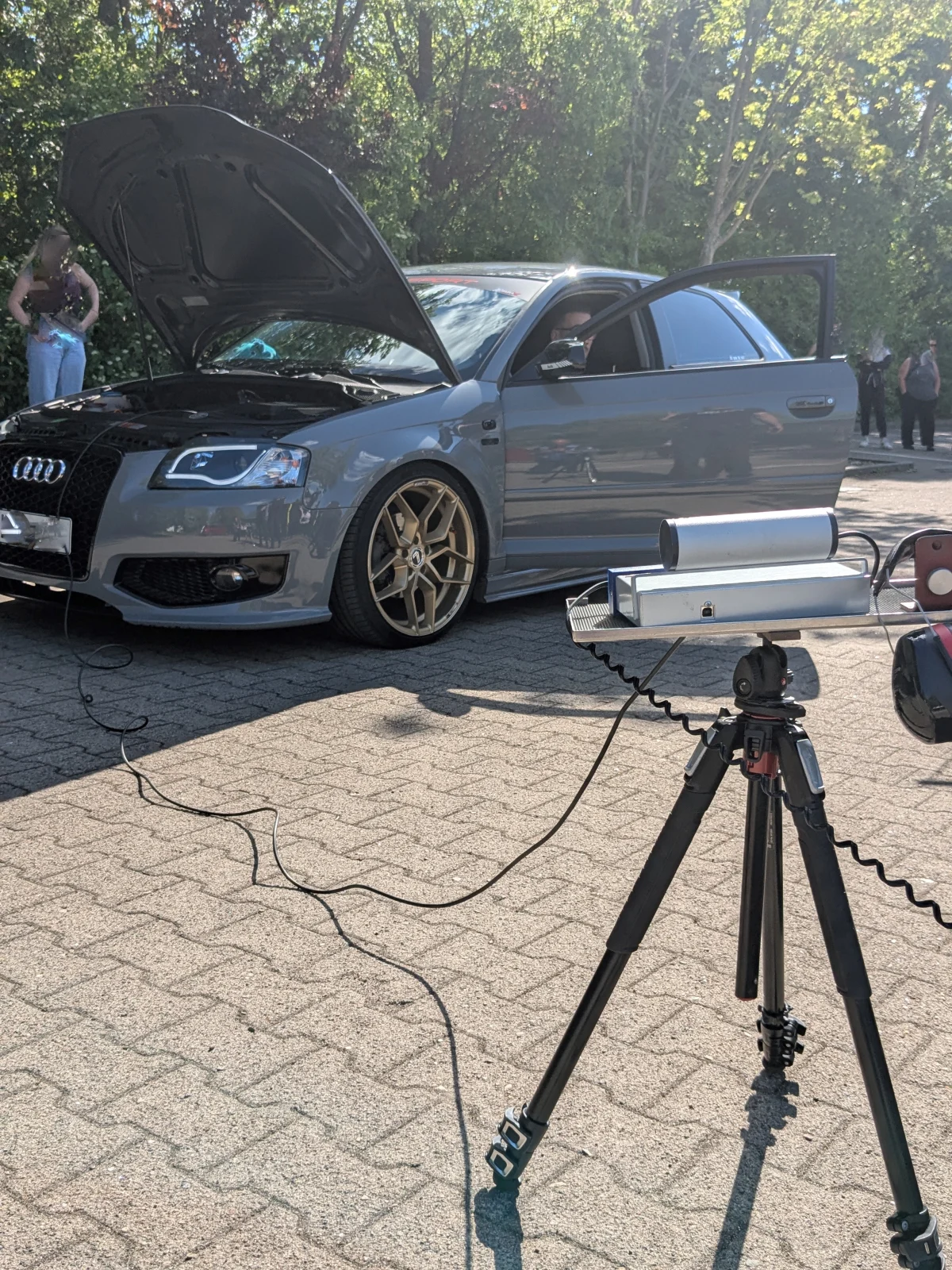 Ein Audi A3 steht mit geöffneter Motorhaube auf einem Parkplatz, während Messgeräte auf einem Stativ daneben platziert sind. Im Hintergrund sind Personen und Bäume erkennbar.