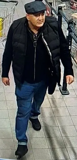 Ein Mann mit dunkler Kleidung und Mütze läuft durch einen Supermarktgang. Im Hintergrund sind Regale und Einkaufswagen sichtbar.