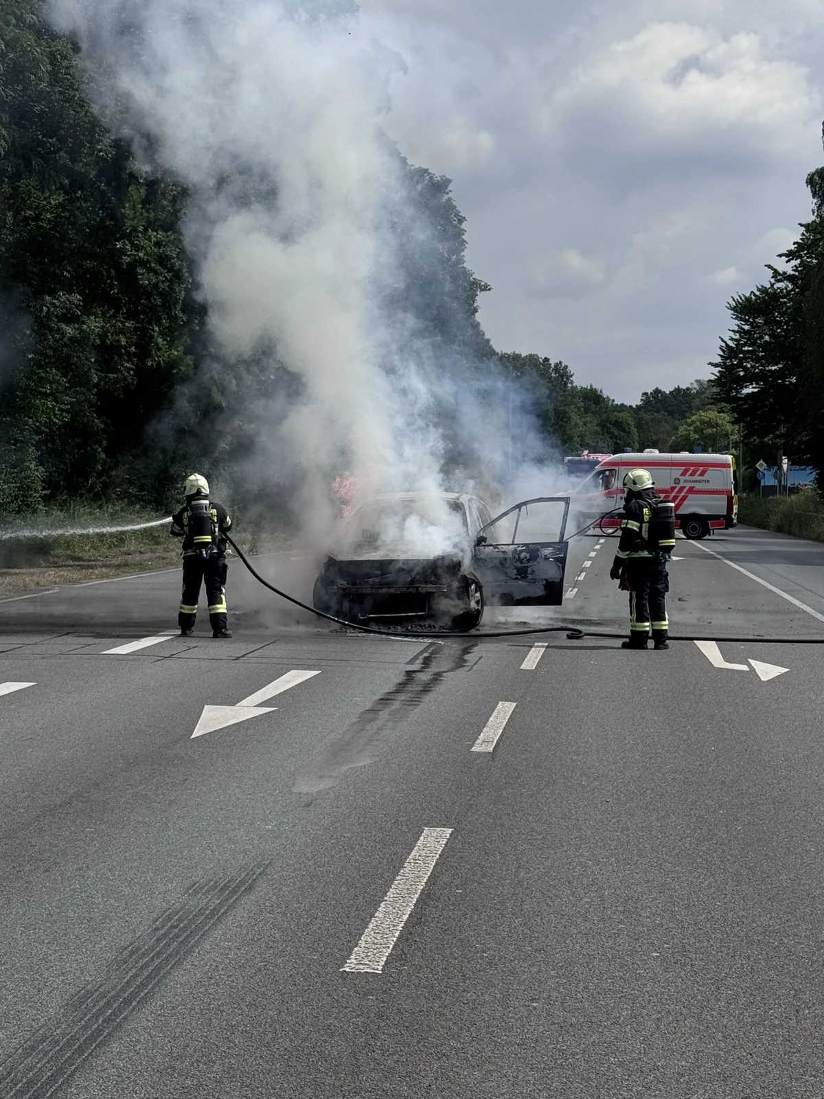 Zwei Feuerwehrleute bekämpfen einen Fahrzeugbrand auf einer Straße, während Rauch aufsteigt. Ein Einsatzfahrzeug steht daneben.