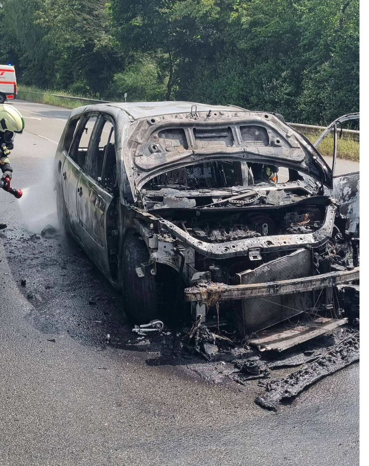 Ein fast völlig ausgebranntes Fahrzeug steht auf einer Straße, während ein Feuerwehrmann die letzten Glutnester löscht. Im Hintergrund ist ein Rettungsfahrzeug sichtbar.