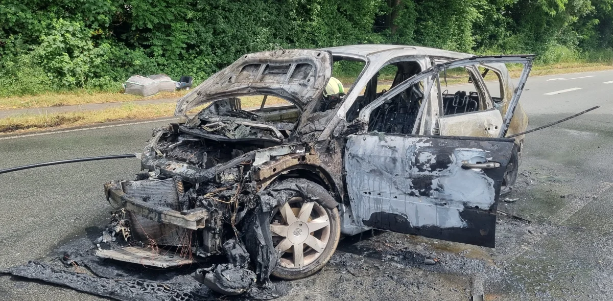 Das Bild zeigt ein ausgebranntes Auto mit geöffneten Türen, das auf einer Landstraße steht. Der Lack ist verbrannt und Teile des Fahrzeugs sind komplett zerstört.