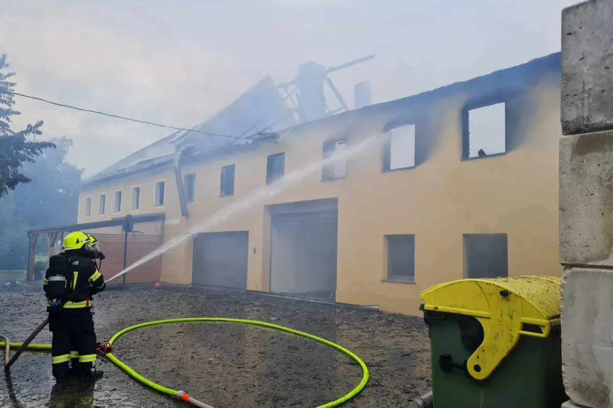Ein Feuerwehrmann in kompletter Schutzausrüstung sprüht Wasser auf ein brennendes, zweistöckiges Gebäude. Rauch steigt von der Dachkonstruktion auf.