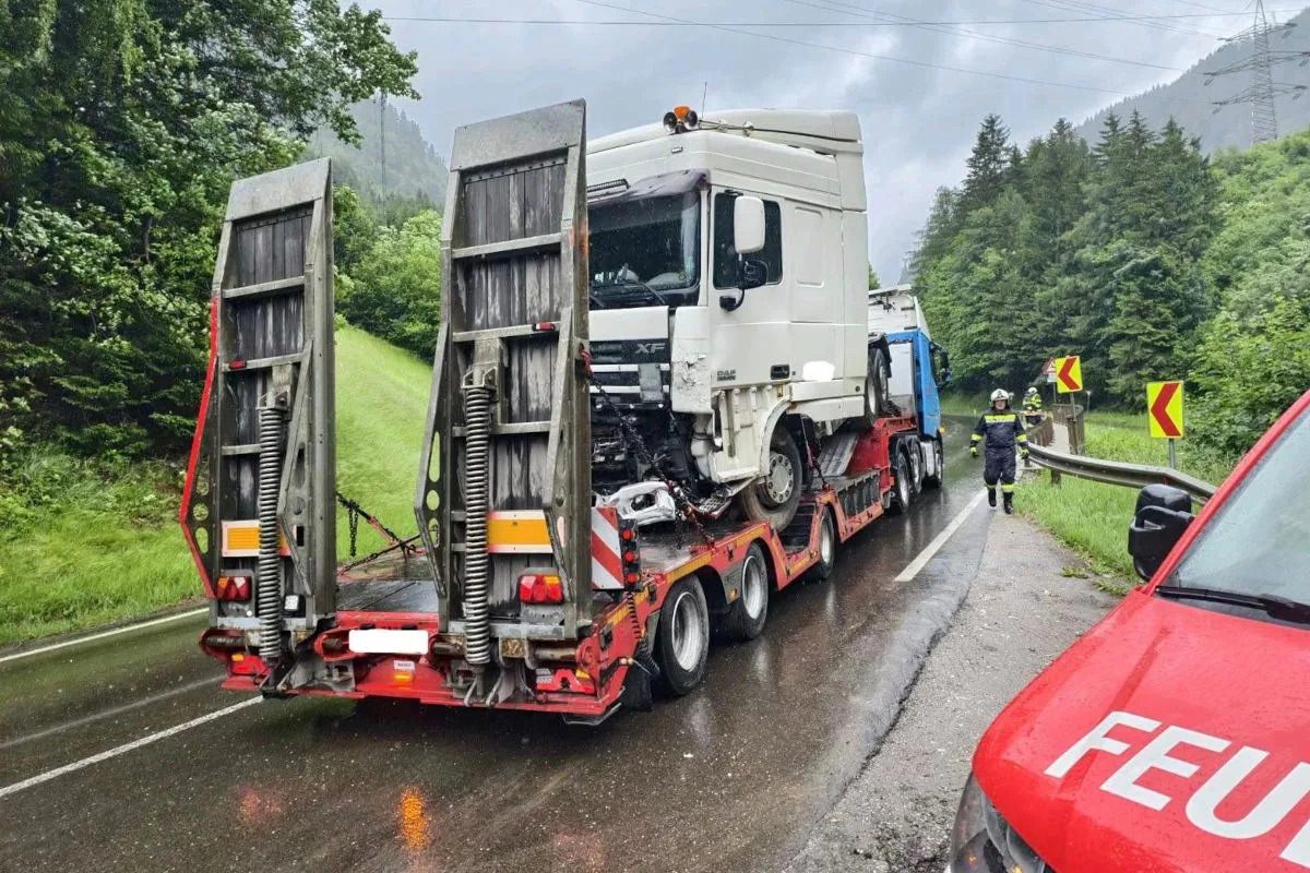 Ein großer, weißer LKW mit beschädigtem Führerhaus wird auf einem roten Anhänger transportiert. Feuerwehrleute sichern die regennasse Straße im Waldgebiet.