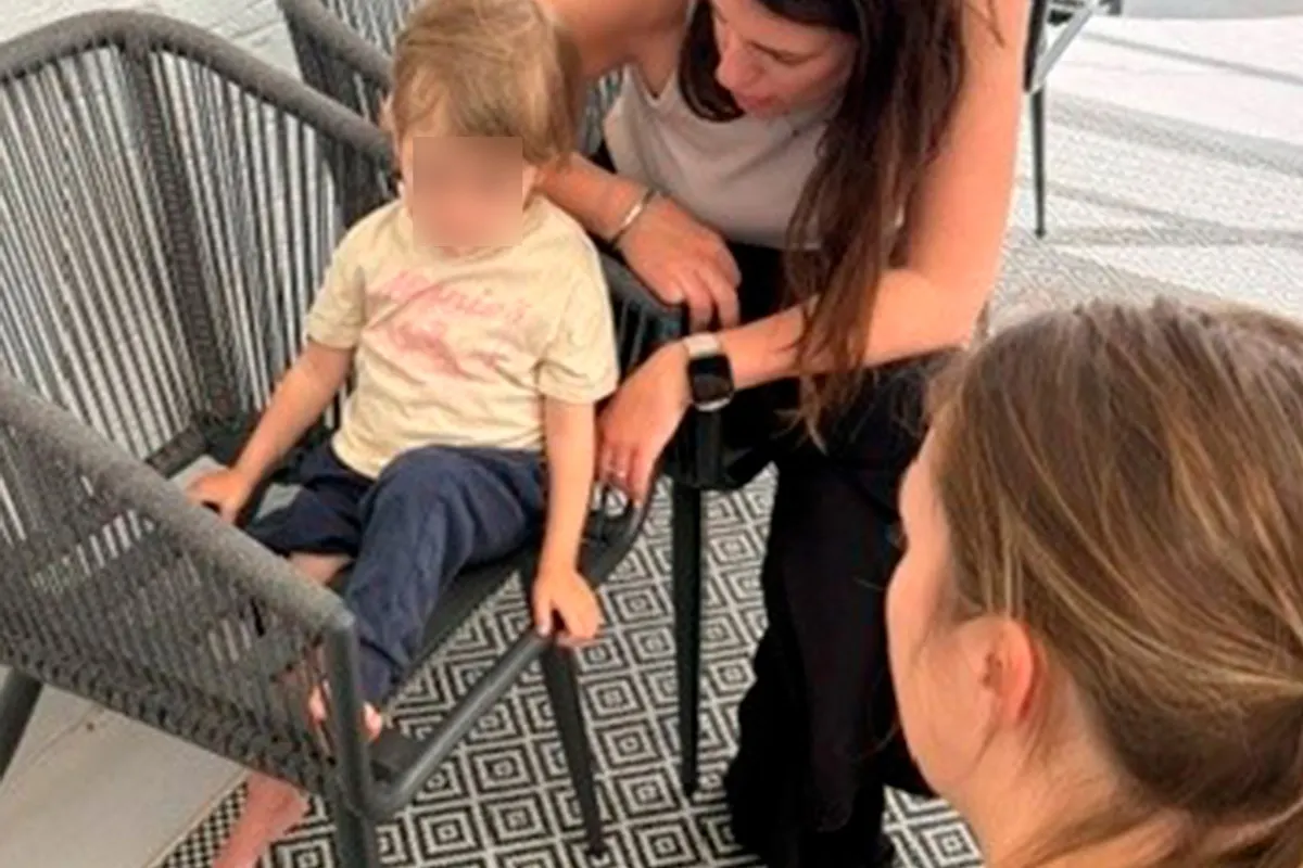 Ein Kind sitzt auf einem Stuhl auf einer Terrasse. Zwei erwachsene Personen lehnen sich vor, um mit dem Kind zu sprechen.