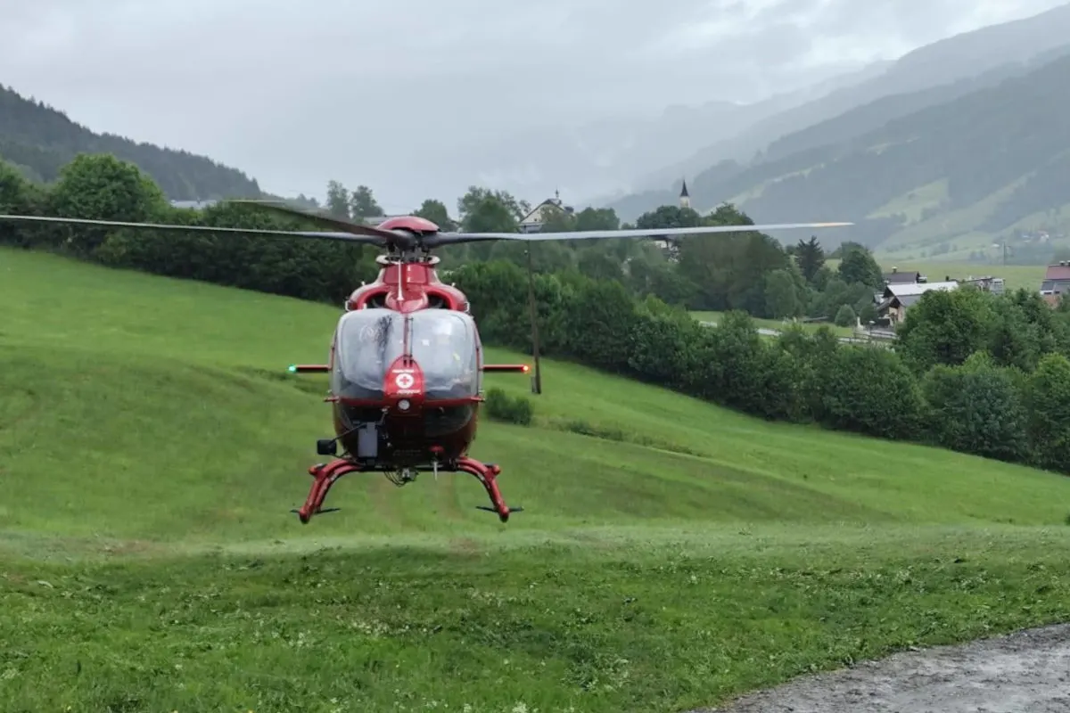 Ein roter Rettungshubschrauber schwebt über einer grünen Wiese in einer bergigen Region. Der Himmel ist bewölkt und eine kleine Siedlung befindet sich im Hintergrund.