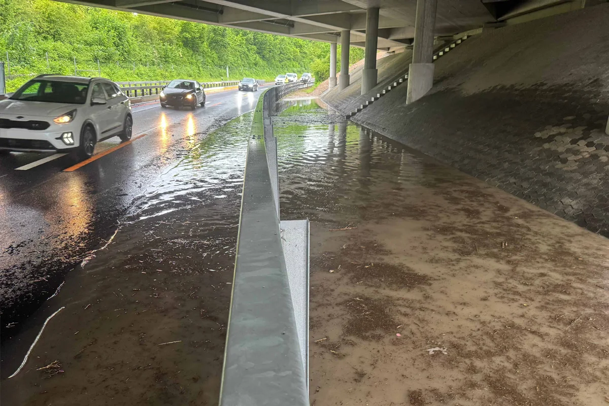 Mehrere Fahrzeuge fahren bei starkem Regen auf einer nassen Straße unter einer Brücke. Rechts davon steht Wasser.