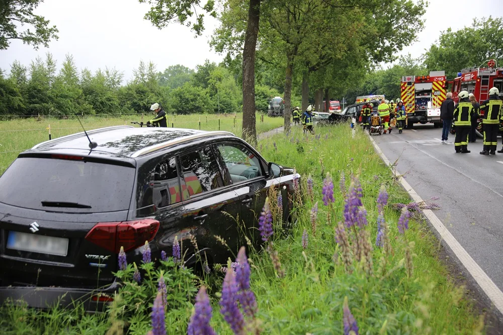 Ein schwarzes SUV steht im Gras am Straßenrand. Dahinter sind Feuerwehrfahrzeuge und Feuerwehrleute bei einem Einsatz zu sehen.