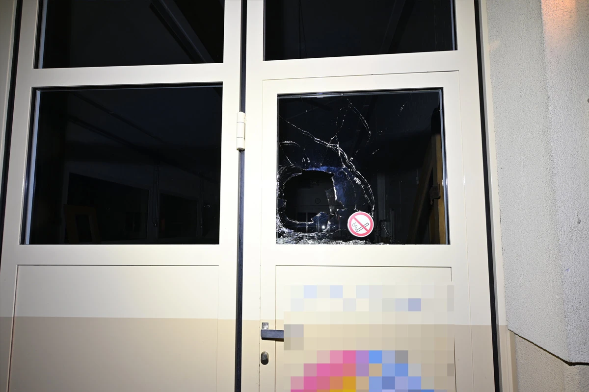 Eine Glastür zeigt ein großes Loch, umgeben von zersplittertem Glas. Ein Schild mit Rauchverbot ist sichtbar.