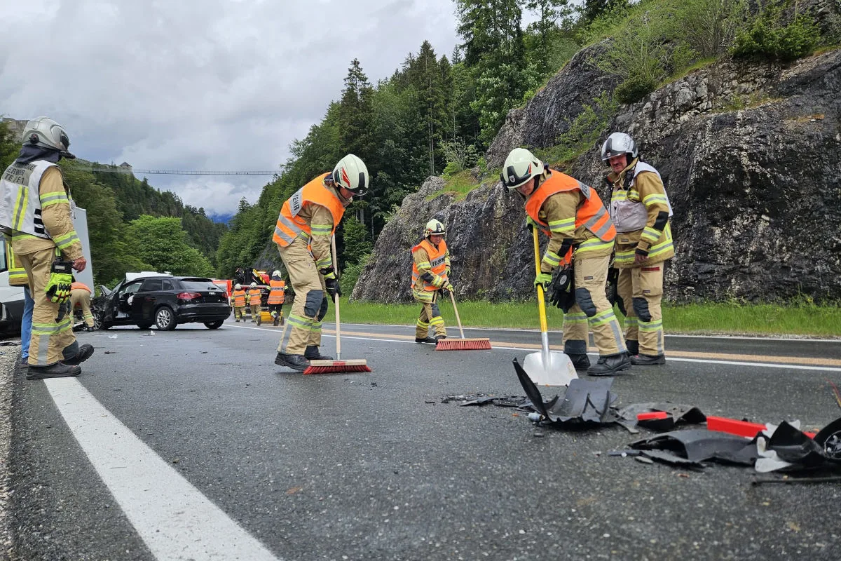 Mehrere Feuerwehrleute in orangefarbenen Westen beseitigen Trümmer auf einer nassen Landstraße nach einem Verkehrsunfall. Im Hintergrund ist ein beschädigtes schwarzes Auto sichtbar.