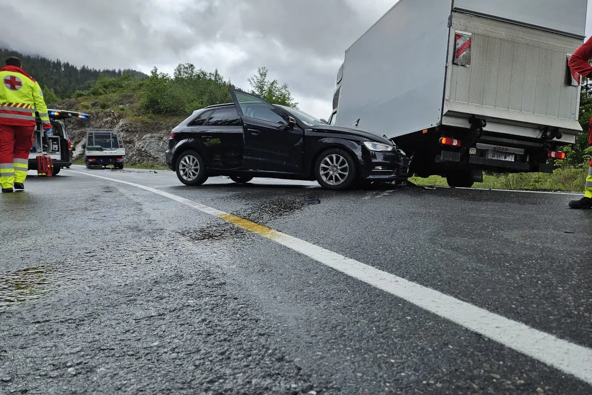 Ein schwarzes Auto hat frontal mit einem weißen Lastwagen auf einer nassen Straße kollidiert. Im Hintergrund sind Rettungskräfte in roten Uniformen zu sehen.