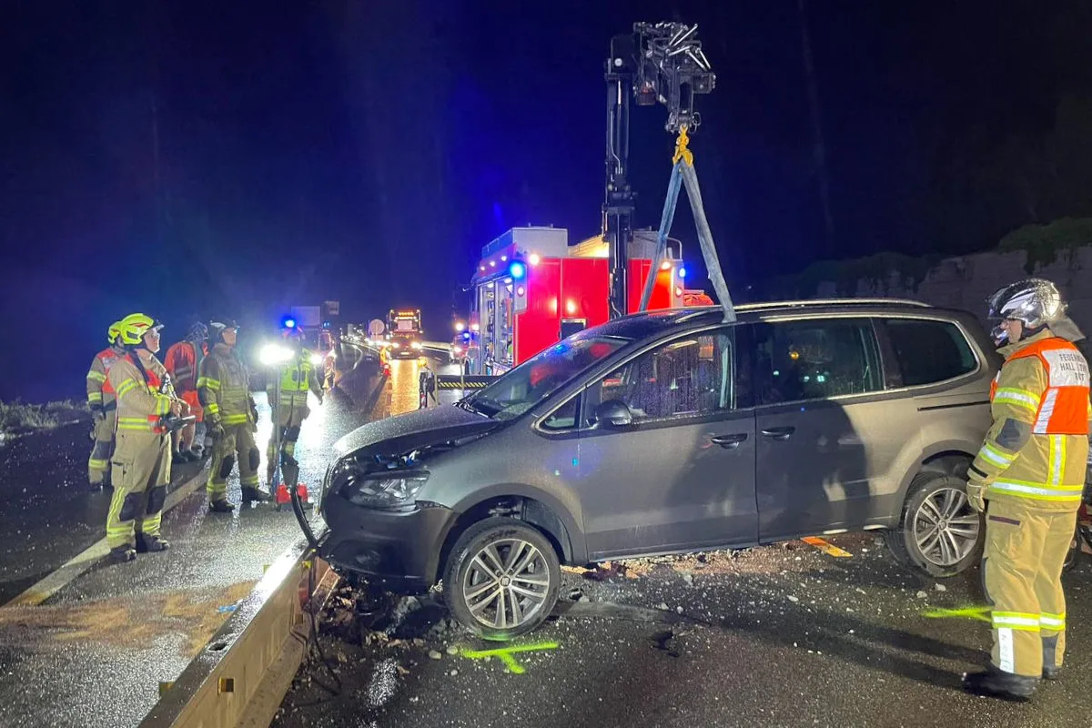 Feuerwehrleute stehen um ein Auto, das von einem Kran angehoben wird. Die Straße ist nass und es ist Nacht.