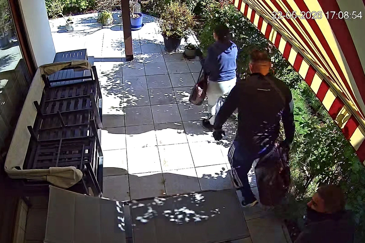 Zwei Personen sind auf einer gefliesten Terrasse unter einer Markise zu sehen, bei Sonnenschein. Eine Person scheint eine Tasche zu tragen.