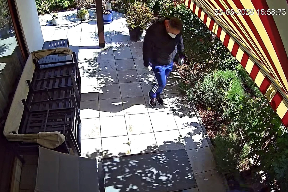 Eine Person mit Maske geht tagsüber auf einer Terrasse entlang. Die Szene ist von Pflanzen und einer Markise umgeben.